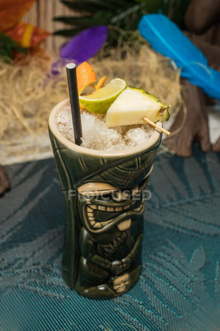 From above of large sculptural tiki cup filled with booze decorated with straw and fruits placed on green rug against dry grass — Stock Photo