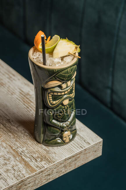 From above of large sculptural tiki cup filled with booze decorated with straw and fruits placed on green rug against wooden table — Stock Photo