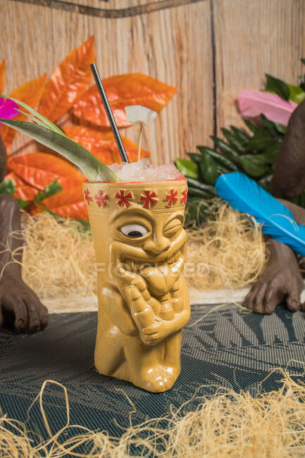 Polynesian tiki cup of cold alcohol beverage decorated with straw and green leaves placed against colorful leaves and dry grass — Stock Photo