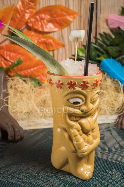 Copa tiki polinesia de bebida alcohólica fría decorada con paja y hojas verdes colocadas contra hojas coloridas y hierba seca - foto de stock