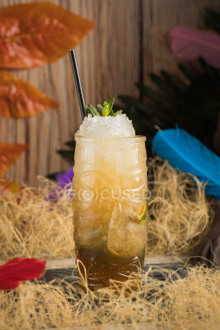 Tiki cup with cold alcohol drink with straw served with ice and decorated with fresh herb placed against dry grass on blurred background — Stock Photo