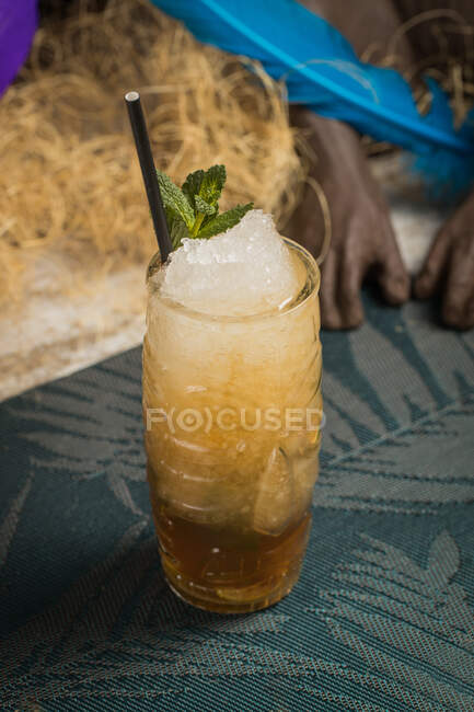 From above of tiki cup with cold alcohol drink with straw served with ice and decorated with fresh herb placed against dry grass on blurred background — Stock Photo