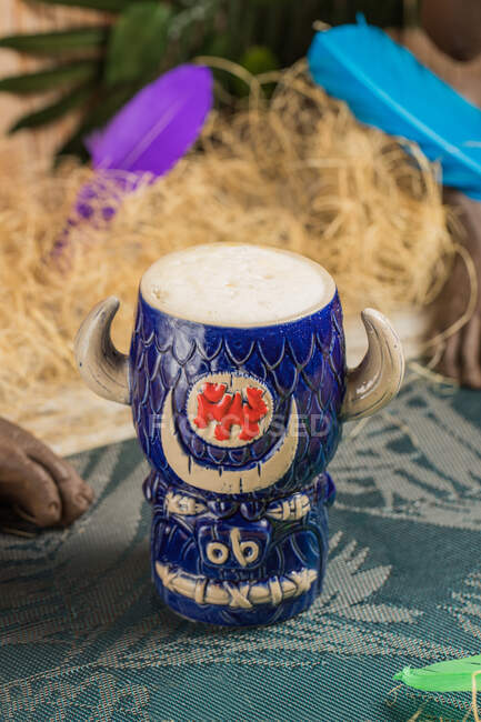 From above of bull shaped tiki mug of alcohol drink with froth placed against dry grass and feathers on blurred background — Stock Photo