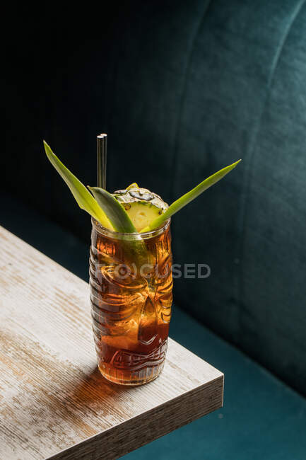 De arriba de la taza de cristal tiki llena de bebida alcohólica con paja decorada con fruta colocada en el borde del sofá de mesa de madera - foto de stock
