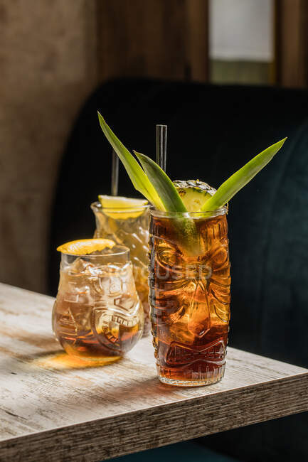 Gläser Tiki-Becher gefüllt mit alkoholischen Getränken mit Stroh verziert mit Früchten auf dem Rand der hölzernen Tischcouch platziert — Stockfoto