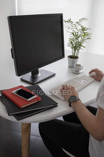 From above of crop unrecognizable male employee in casual clothes and wristwatch typing on computer keyboard while working at table with notebooks and potted plant in modern office — Stock Photo