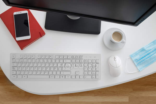 Moderner Computer und Smartphone mit Notizbuch auf weißem Tisch mit medizinischer Maske im hellen Büro — Stockfoto