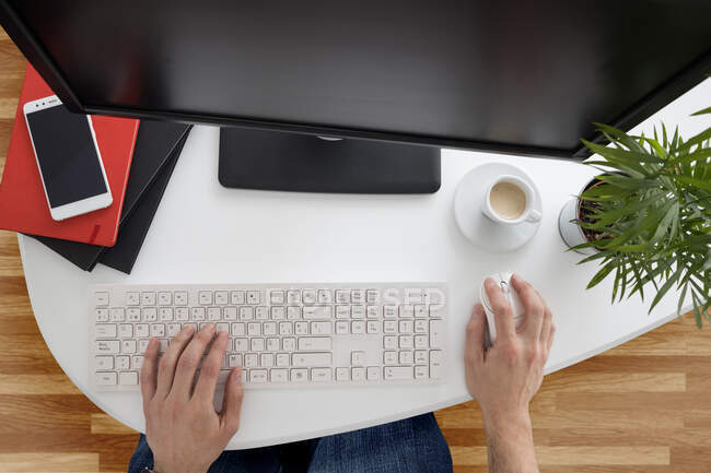 Вид сверху неузнаваемого сотрудника-мужчины в повседневной одежде и наручных часах, сидящего на клавиатуре компьютера, работающего за столом с ноутбуками и горшочком в современном офисе — стоковое фото