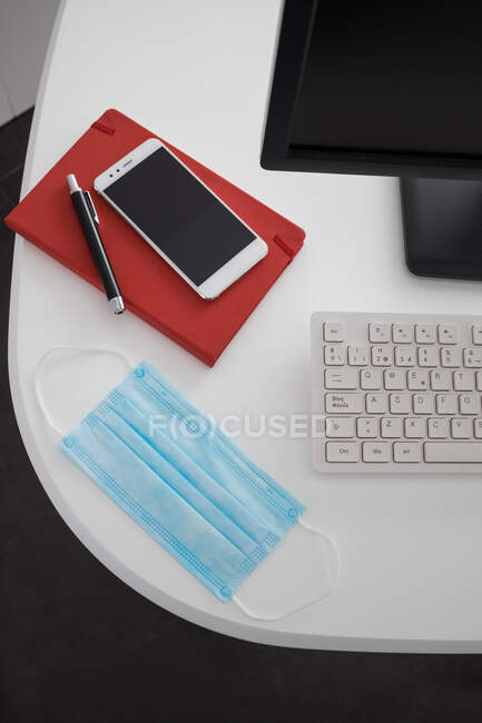 Зверху сучасний комп'ютер і смартфон з блокнотом, розміщеним на білому столі з медичною маскою в легкому офісі — стокове фото