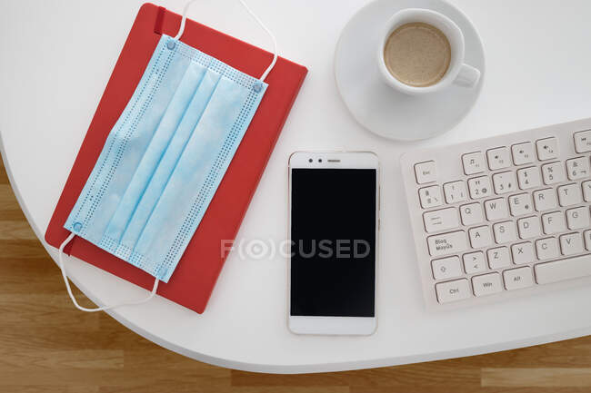 Vista dall'alto della maschera medica posta sul taccuino e disposta con smartphone e tastiera vicino alla tazza di caffè sulla scrivania bianca — Foto stock