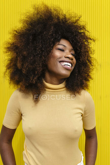Jeune femme ethnique joyeuse avec coiffure afro et les yeux fermés riant sur fond jaune — Photo de stock