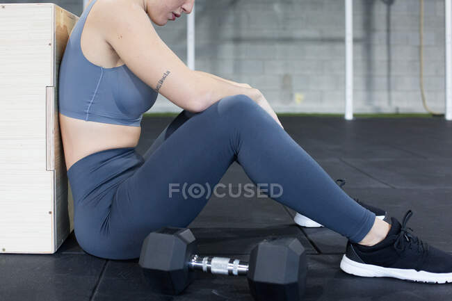Side view of crop unrecognizable exhausted female athlete sitting on floor near dumbbells and resting after intense functional workout in sports center — Stock Photo