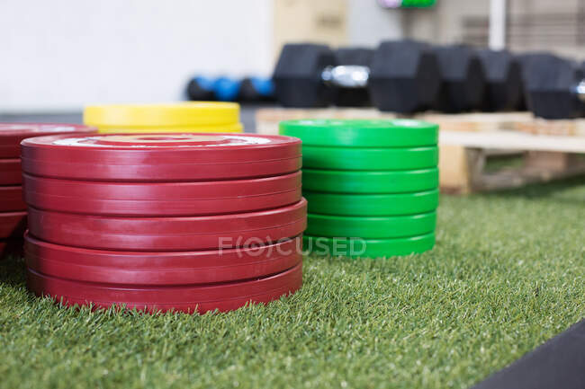 Placas multicoloridas de peso de metal pesado empilhadas na grama artificial no ginásio moderno com vários equipamentos esportivos — Fotografia de Stock