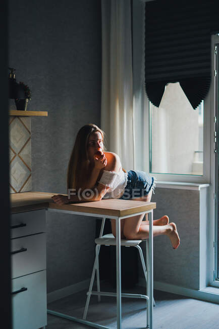 Приваблива мрійлива жінка в білому верху з голими плечима, що тримає солодкий персик і дивиться в сторону, спираючись на кухонну стійку — стокове фото
