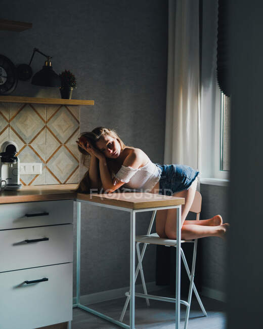Attractive dreamy female in white top with bare shoulders and looking at camera while leaning on kitchen counter — Stock Photo