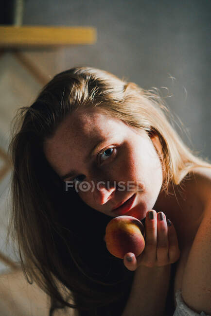 Mulher sonhadora atraente em top branco com ombros nus segurando pêssego doce e olhando para a câmera enquanto se inclina no balcão da cozinha — Fotografia de Stock