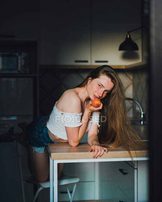 Attraktive verträumte Frau in weißem Top mit nackten Schultern, die süßen Pfirsich in der Hand hält und in die Kamera schaut, während sie sich auf den Küchentisch lehnt — Stockfoto
