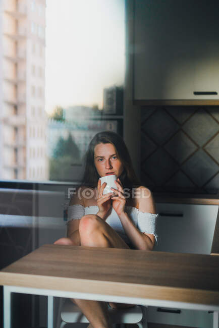 Молода жінка з довгим волоссям носить повсякденний одяг з голими плечима, насолоджуючись свіжим гарячим напоєм і дивлячись на камеру, спираючись на кухонну стійку — стокове фото