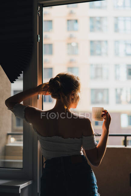 Back view serene young female with long hair wearing casual clothes with bare shoulders enjoying fresh hot drink and looking away on kitchen — Stock Photo
