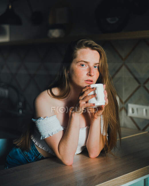 Serene young female with long hair wearing casual clothes with bare shoulders enjoying fresh hot drink and looking at camera while leaning on kitchen counter — Stock Photo
