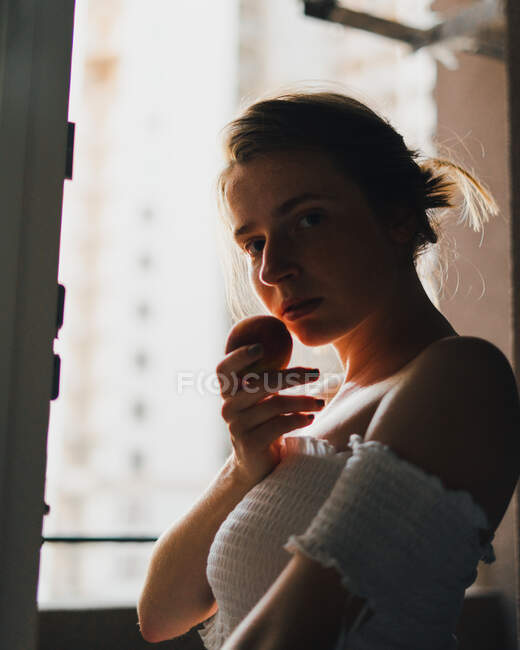 Attractive dreamy female in white top with bare shoulders holding sweet peach and looking at camera — Stock Photo