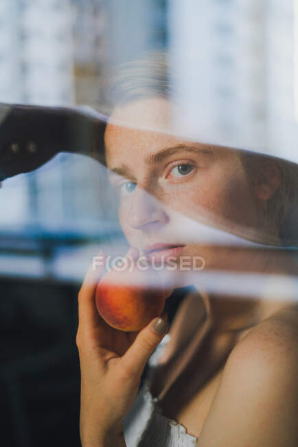 Attraktive verträumte Frau in weißem Top mit nackten Schultern, die süßen Pfirsich in der Hand halten und durch das Glas in die Kamera schauen — Stockfoto