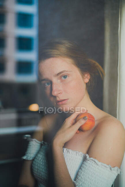 Attraktive verträumte Frau in weißem Top mit nackten Schultern, die süßen Pfirsich in der Hand halten und durch das Glas in die Kamera schauen — Stockfoto