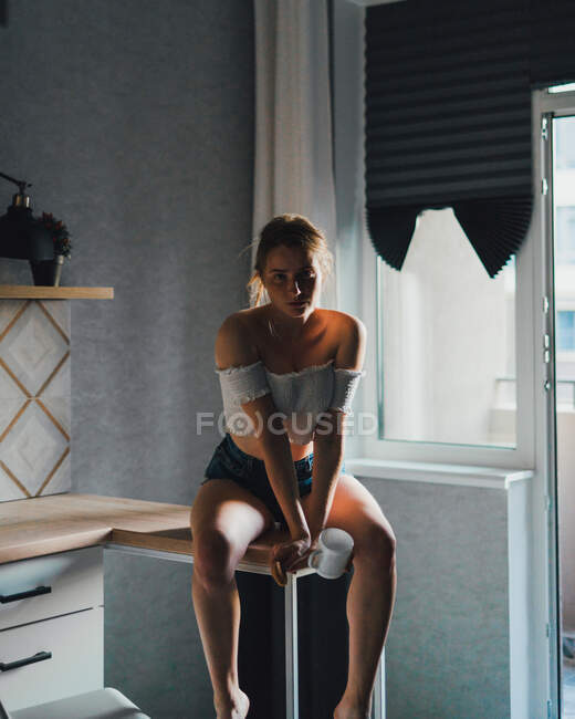 Unemotional young female in shorts and top with bare shoulders sitting with cup of drink on kitchen counter and looking at camera calmly — Stock Photo