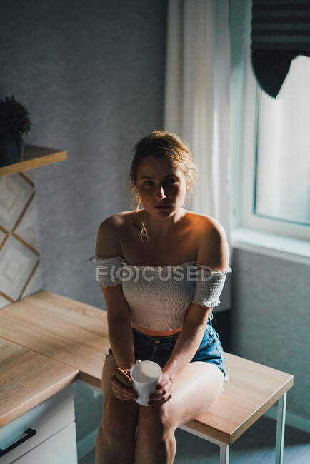 Неэмоциональная молодая женщина в шортах и топе с голыми плечами сидит с чашкой напитка на кухонном столе и спокойно смотрит в камеру — стоковое фото