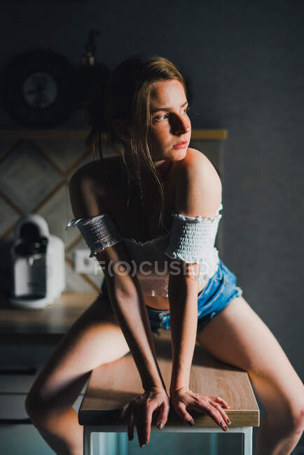 Неэмоциональная молодая женщина в шортах и топе с голыми плечами сидит на кухонном столе и спокойно смотрит в сторону — стоковое фото