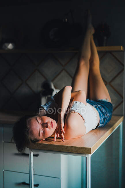 Giovane femmina con gli occhi chiusi in pantaloncini e top con spalle nude sdraiato sul bancone della cucina — Foto stock
