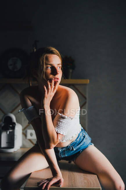 Mujer joven sin emociones en pantalones cortos y la parte superior con hombros desnudos sentado en el mostrador de la cocina y mirando hacia otro lado con calma - foto de stock