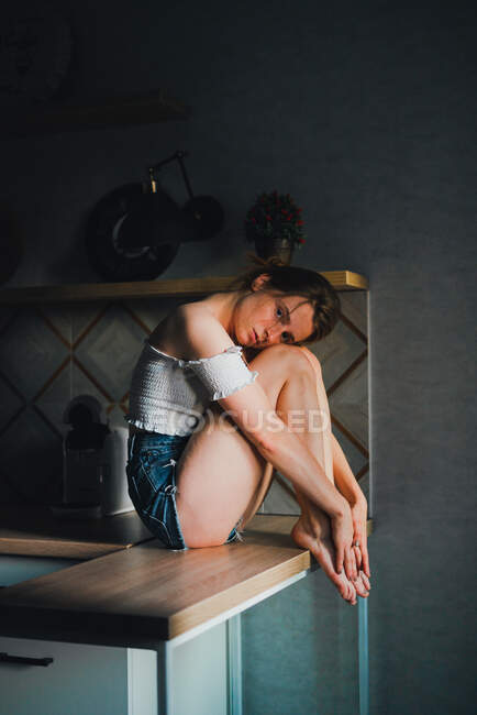 Unemotional young female in shorts and top with bare shoulders sitting on kitchen counter and looking at camera calmly — Stock Photo
