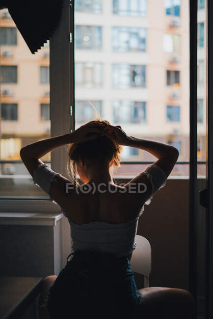 Анонимный вид сзади подходит женщине в повседневной одежде, поднимающей руки и прижимающей волосы к стене дома — стоковое фото