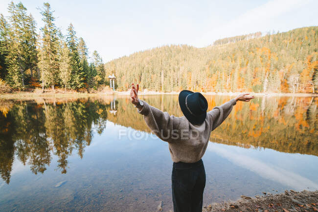 Вид ззаду анонімного туриста-жінки в капелюсі з піднятими обіймами, що милуються прозорою водою проти осінніх дерев на горі — стокове фото