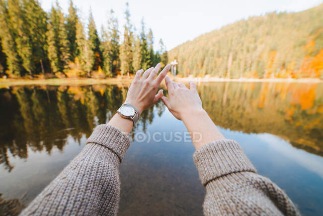 Crop anonymous female tourist in knitted wear with raised arms against pure water and trees on mountain during trip — Stock Photo