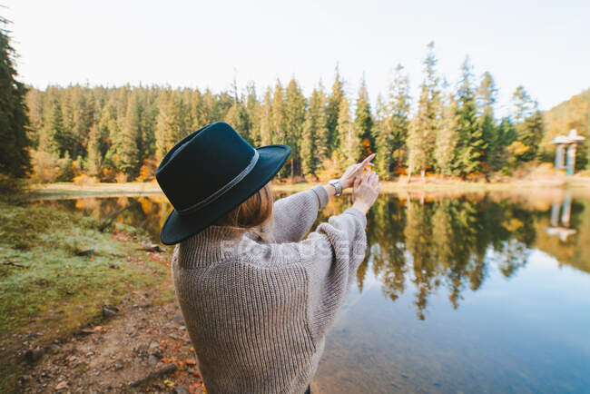 Back view anonymous female tourist in knitted wear with raised arms against pure water and trees on mountain during trip — Stock Photo
