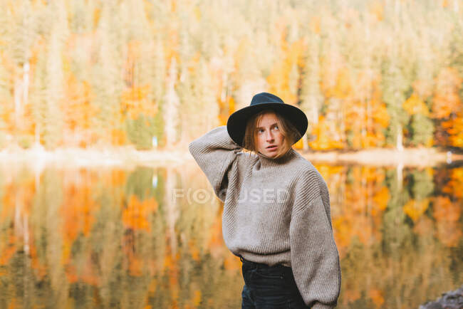Joven turista rubia fresca con ropa de moda mirando hacia otro lado mientras está de pie en la costa contra el agua reflejando árboles - foto de stock