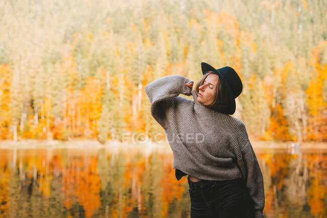 Young blond cool female tourist in trendy apparel with eyes closed standing on coast against water reflecting trees — Stock Photo