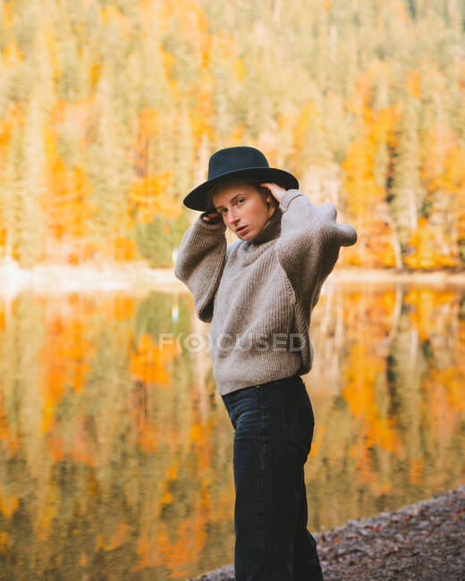 Vista lateral da jovem loira fresca turista feminina em vestuário da moda olhando para a câmera enquanto está em pé na costa contra a água refletindo árvores — Fotografia de Stock