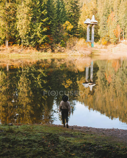 Back view of anonymous female tourist in hat standing admiring transparent water against autumn trees on mount — Stock Photo