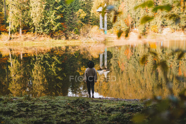 Back view of anonymous female tourist in hat standing admiring transparent water against autumn trees on mount — Stock Photo