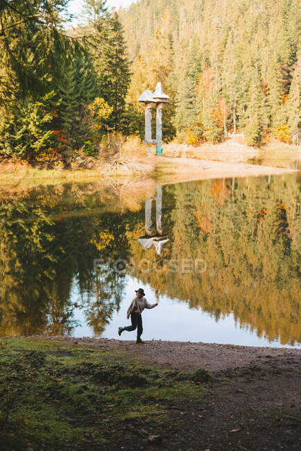 Anonymous female tourist with raised arm and leg running on shore against water reflecting coniferous trees and looking away — Stock Photo