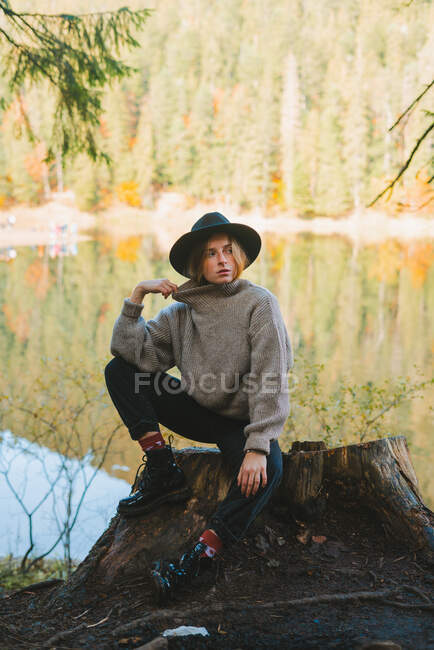 Молода модна жінка-туристка в капелюсі торкається обличчя, дивлячись в сторону і відпочиваючи проти яскравих дерев, що відображаються у воді — стокове фото