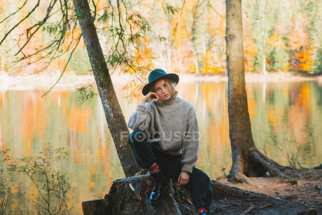 Young trendy female tourist in hat touching face while looking at camera and resting against bright trees reflecting in water — Stock Photo