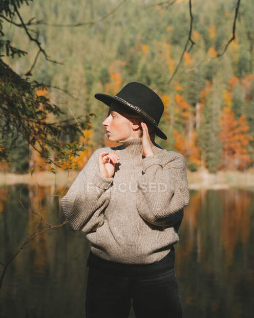 Joven turista rubia fresca con ropa de moda mirando hacia otro lado mientras está de pie en la costa contra el agua reflejando árboles - foto de stock