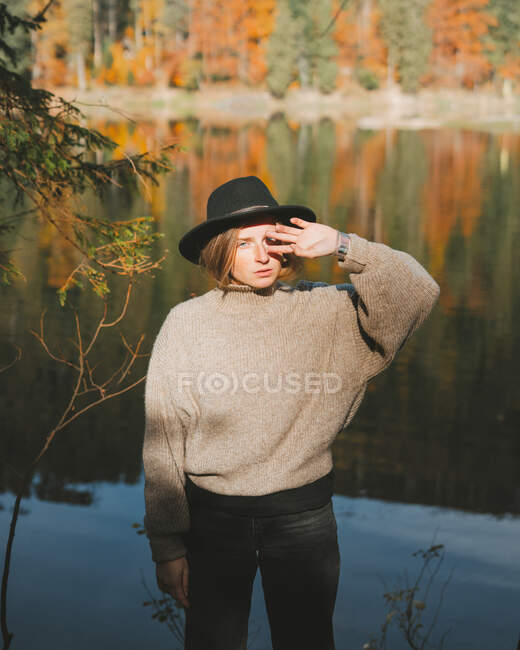 Молодая блондинка прохладный женщина турист в модной одежде глядя в камеру, стоя покрывая лицо на берегу против воды отражая деревья — стоковое фото