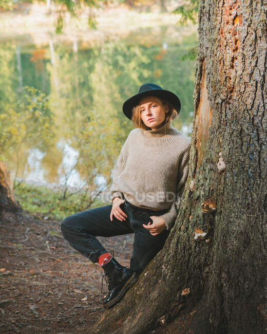 Young trendy female tourist in hat looking at camera while resting standing leaning on tree near lake near autumn — Stock Photo
