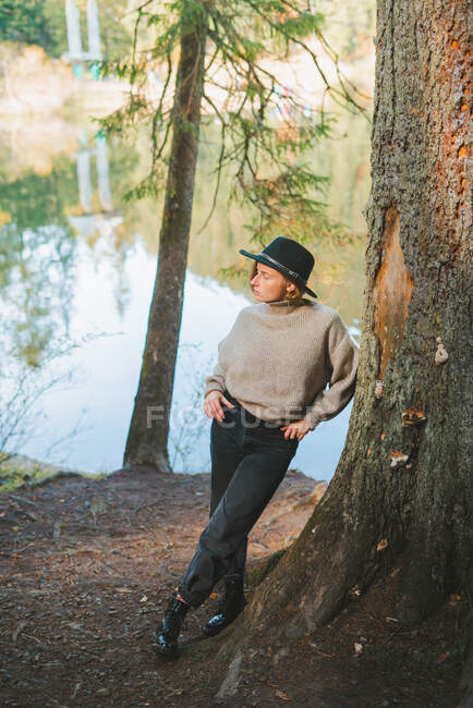 Jovem turista na moda em chapéu olhando para longe enquanto descansa em pé apoiando-se na árvore perto do lago perto do outono — Fotografia de Stock