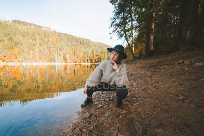 Молодая крутая туристка в модной одежде смотрит вдаль от сухого берега на прозрачную воду, отражающую осенние деревья — стоковое фото